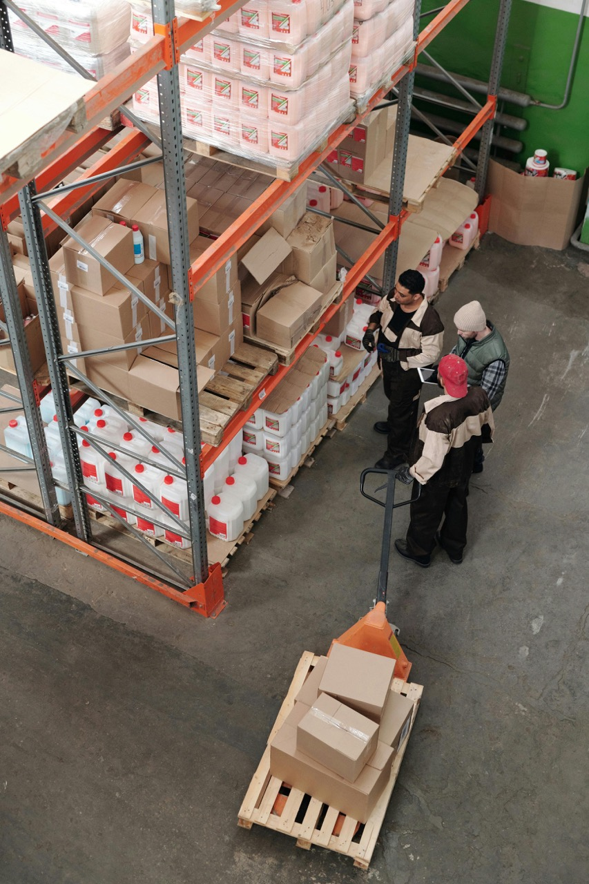 Workers in a warehouse organizing and inspecting boxes on a pallet, surrounded by shelves stocked with various containers and packages