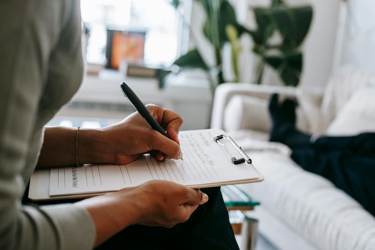 Close-up of a person writing on a clipboard while another person relaxes on a couch in the background, suggesting a therapy or counseling session.