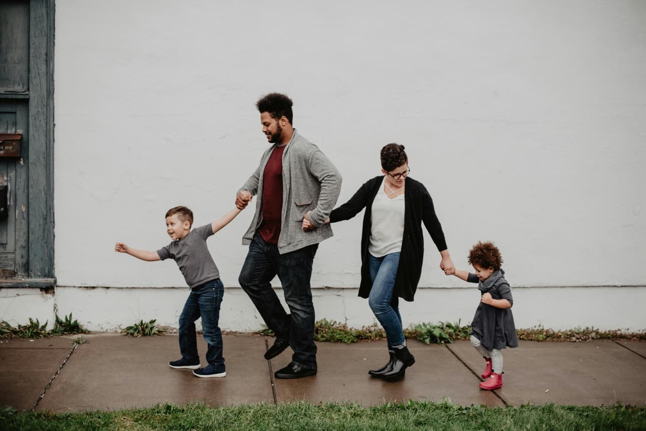 A family of four walking hand in hand on a sidewalk, with a young boy leading, followed by his father, mother, and a younger girl at the end