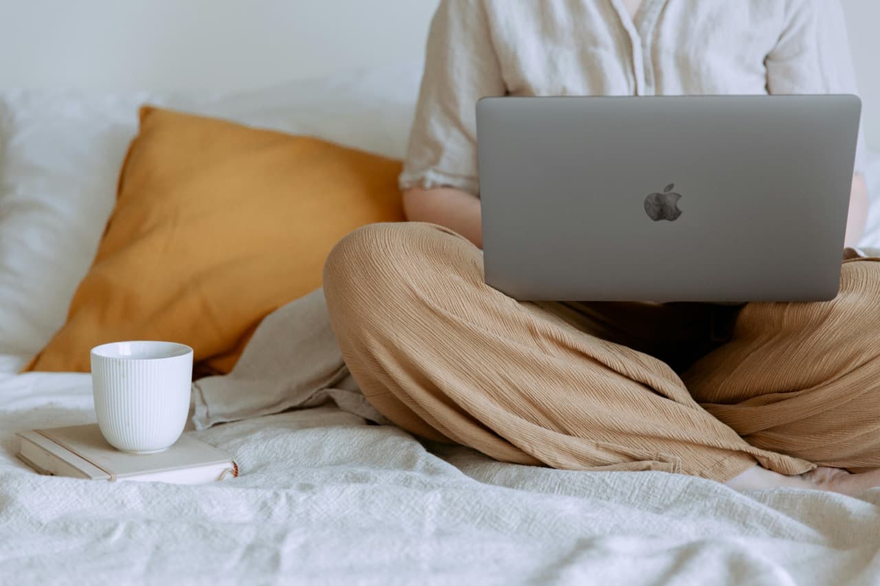 A person is using Macbook on a bed with coffee on the side